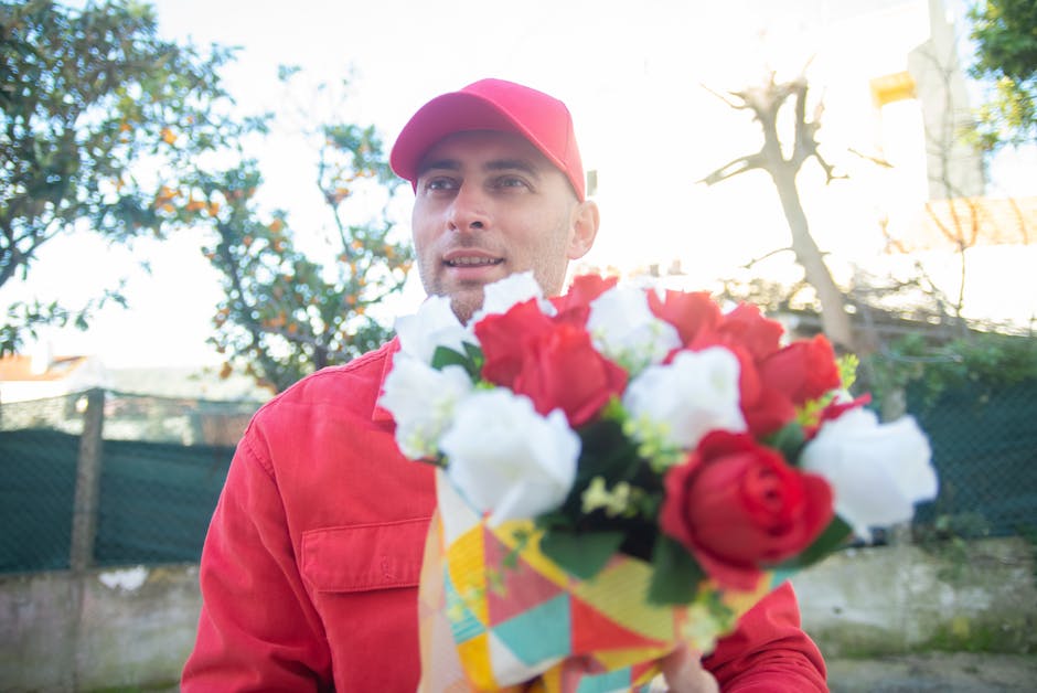 A man dressed in a red jacket and matching red cap is holding a floral bouquet with red, white, and green flowers, wrapped in colorful paper. He is standing outdoors with trees and a fence in the back