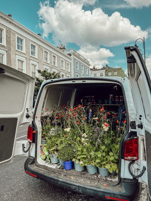 The open rear of a white delivery van filled with a variety of potted flowers and blooming plants, including red, white, and green foliage, arranged in blue and gray pots. The flowers are freshly pick