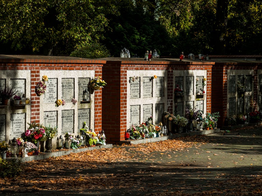A series of memorial plaques mounted on a brick wall in Nunhead Cemetery, adorned with various floral tributes. The flowers include vibrant pink roses, white lilies, purple chrysanthemums, and yellow 