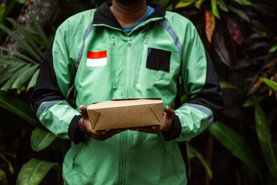 A person wearing a green delivery jacket with a Netherlands flag patch and black accents is holding a rectangular brown paper package, standing amidst lush green tropical foliage. The person’s face 