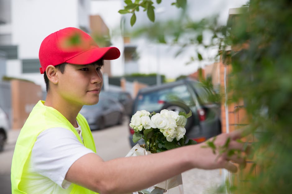 A young delivery person wearing a red cap and a neon yellow safety vest is placing a bouquet of white hydrangeas wrapped in brown paper against a leafy green fence on a residential street. The flowers