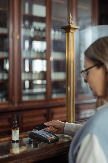 A woman with glasses and blonde hair, dressed in a light-colored blouse, is placing a credit card into a card reader on a wooden counter. In front of her, a small amber glass bottle with a dropper cap